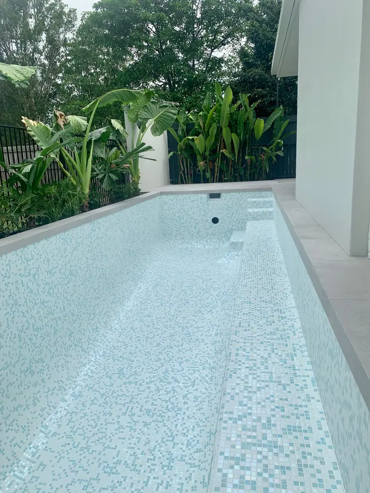 Completed lap pool in white and aqua mosaic tiles beside a tropical garden